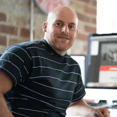 Joe Savery, wearing a dark green horizontally striped shirt, sits smiling in front of a computer monitor in a brick-walled room.