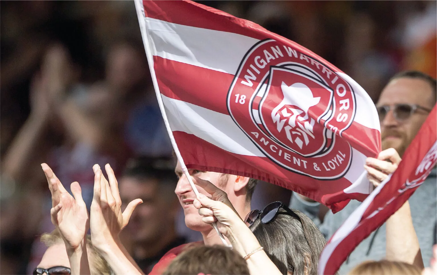A Wigan Warriors flag is flown in the stands of a rugby match by a fan.