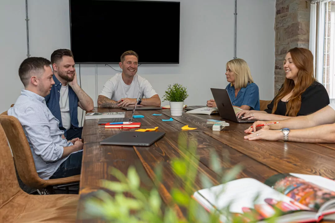Five people sit around a long wooden table, working on laptops and papers. They are engaged in a meeting in a modern office setting.