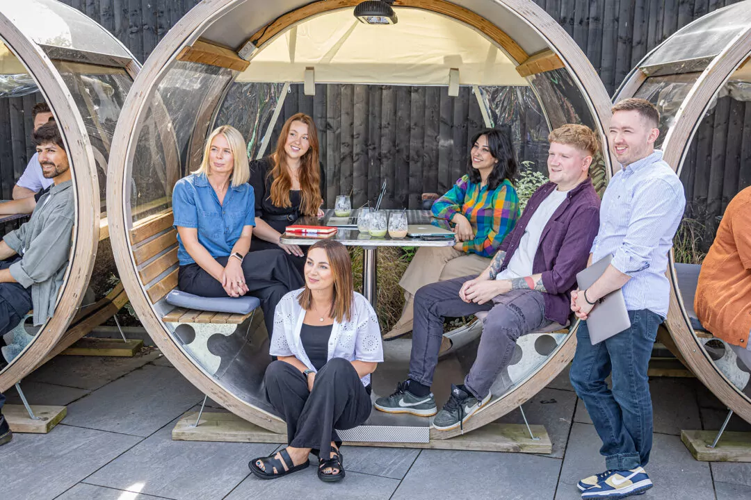 Eight people sit and stand in semi-circular wooden booths outdoors. They appear to be in a casual meeting, with laptops and drinks present.