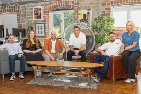 Six people, seated on a grey couch and brown leather chair, pose for a photograph in a brick-walled room. A low coffee table sits before them.