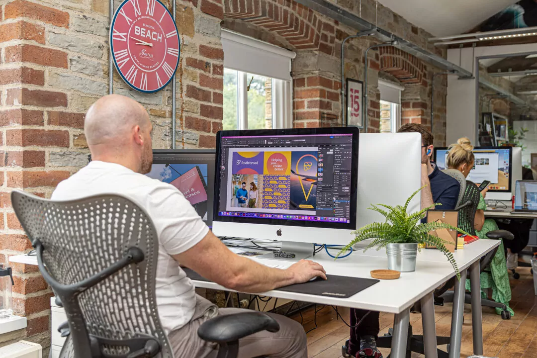 Joe designs marketing materials using a computer in a brick-walled office. Two other coworkers work nearby.