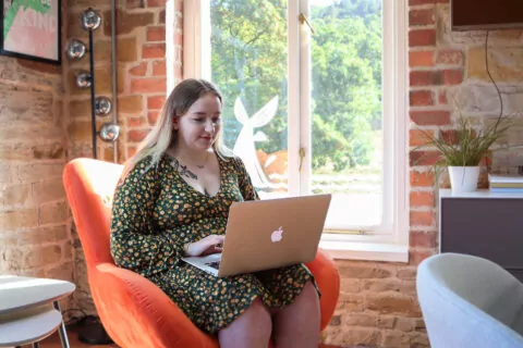 Chloe uses a laptop in an orange armchair. She sits near a large window in a room with exposed brick walls.