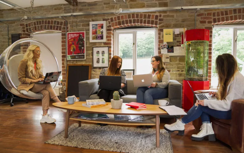 Four women work on laptops in a rustic, brick-walled office. One sits in a transparent chair, the others on a sofa. A large fish tank stands nearby.