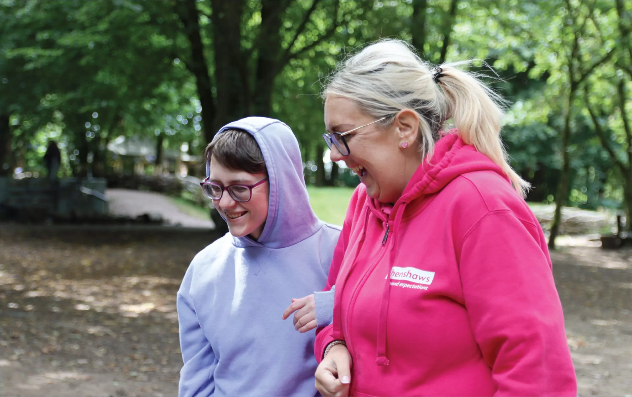 Two people are in a wooded area, one wears a grey hoodie, while the other wears a branded 'Henshaws' hoodie in pink.