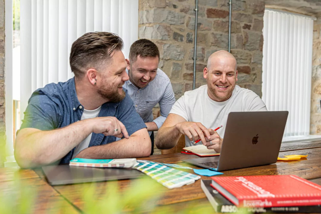 Mike, Tom and Joe collaborate, using a laptop and colour swatches; they are seated at a wooden table in a brightly lit room.