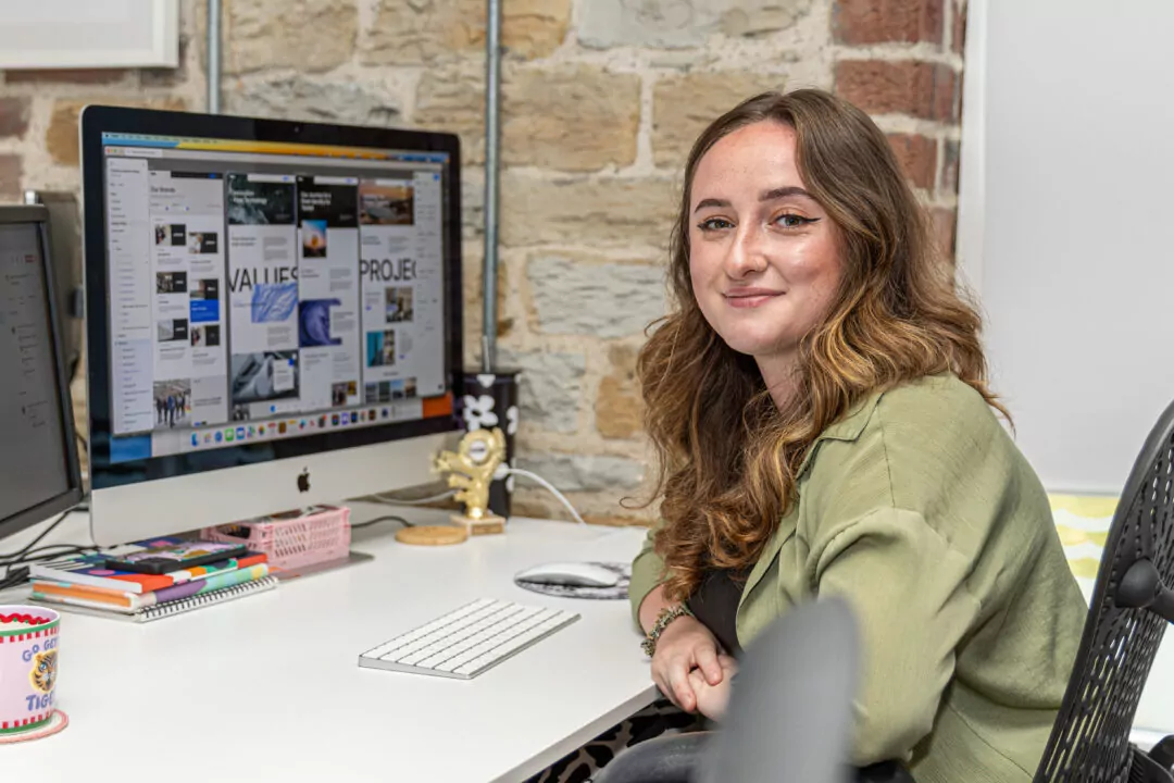 Emily sat at a desk, smiling at the camera. She is working on a computer displaying various webpage designs.