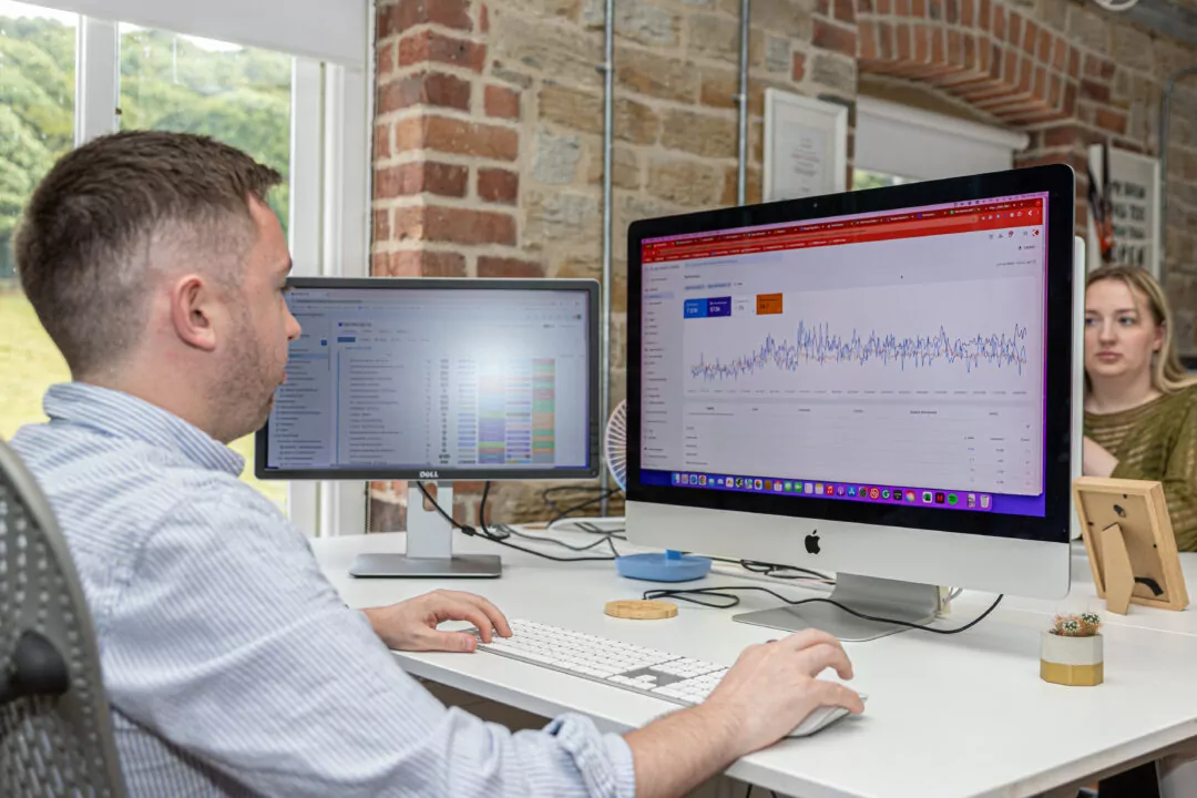 Tom uses a computer keyboard and mouse, analysing data displayed on two monitors in an office. Chloe sits in the background.