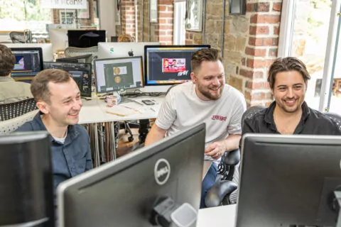 Paul, Mike and Charlie collaborate, smiling at computer screens in a modern office.