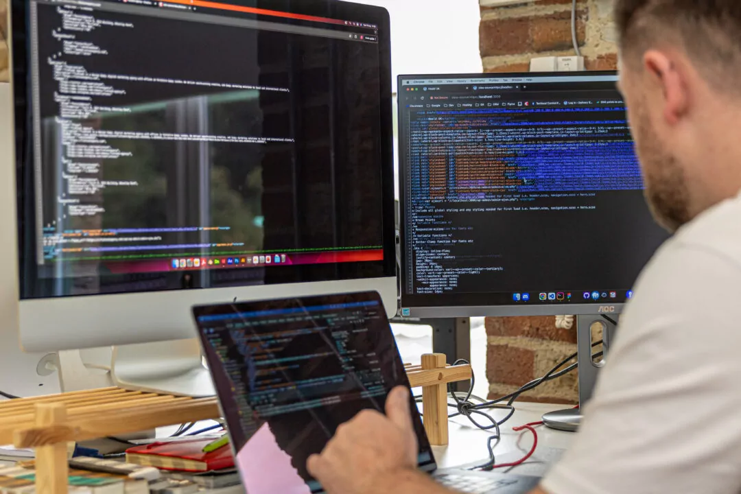 A person reviews lines of code displayed on three screens: a large desktop monitor, a smaller secondary monitor, and a laptop. The screens show various programming languages. The context is a workspace with brick walls.