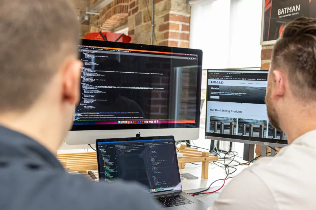 Two programmers work on computer code, displayed on desktop and laptop screens, HEALD appears on one screen.