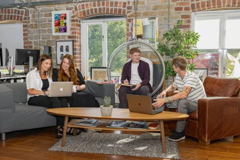Four individuals work collaboratively in a loft-style office. Two women use laptops on a couch; a man in a hanging chair takes notes; another man interacts with them.