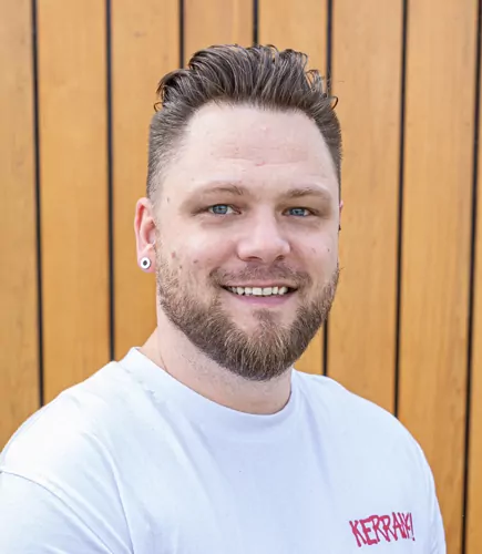 Mike smiles, wearing a white t-shirt with "KERRANG!" printed on it, standing in front of a light brown wood-panelled wall.