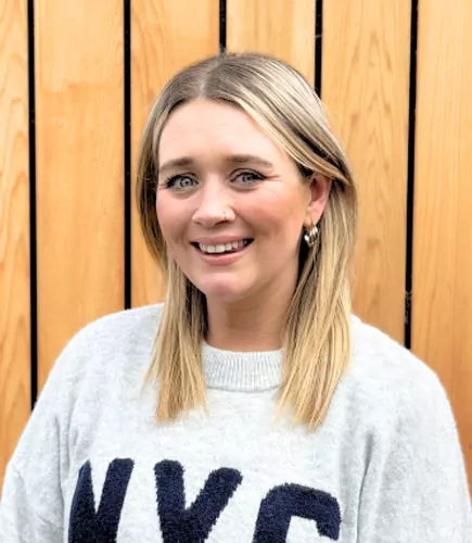 Jess smiles, wearing a light grey sweater with “NYC” on it, in front of a vertical wooden wall.