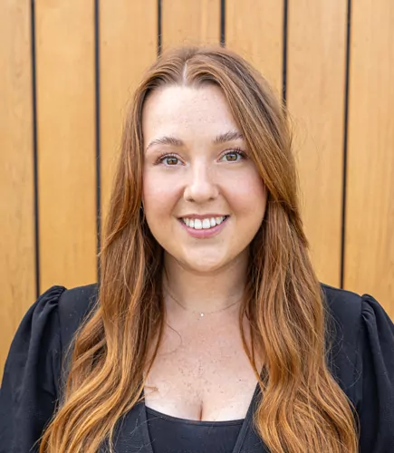 Jacy, a woman with long, auburn hair smiles while wearing a black top; she is standing in front of a light brown wood plank wall.