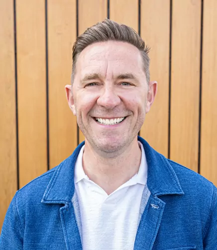 Damien smiles broadly. He wears a blue jacket over a white collared shirt. The background is a light brown vertical-panelled wall.