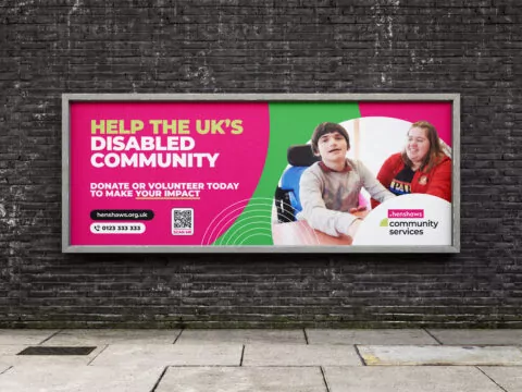 A billboard displays a vibrant advertisement advocating for the UK's disabled community, urging donations or volunteering. Two people, one in a wheelchair, are featured. The billboard is mounted on a brick wall outdoors.
