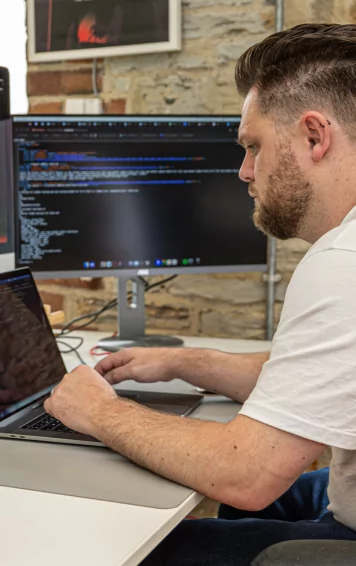 Mike, head of the web development team, types on a laptop, positioned before a large monitor displaying code. He sits at a white desk in a room with a stone wall.