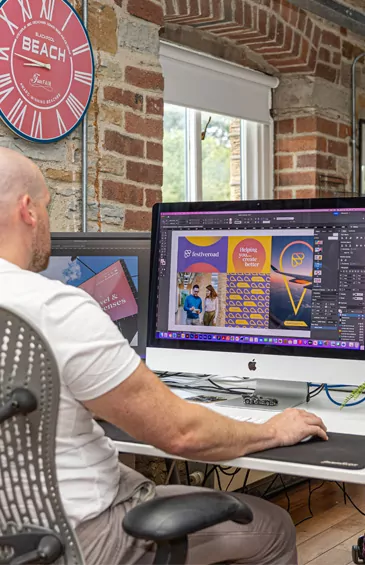 Joe uses a computer to design vibrant creative branding graphics; he sits at a desk in a brick-walled room.