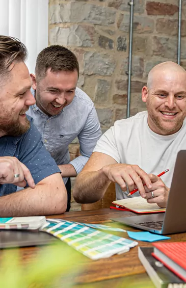 Three men collaborate, laughing while reviewing a laptop and colour swatches at a wooden table in a casual office setting.