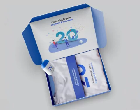 A blue box contains a white t-shirt, a small bottle, and a blue insert. The box's interior and insert celebrate a company's 20th anniversary.
