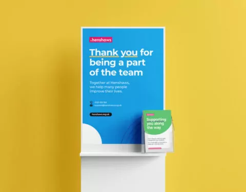 A poster displays a thank you message to team members, alongside a smaller leaflet offering support. Both are displayed on a white stand against a yellow wall.