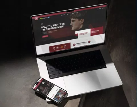 A laptop displays a website featuring a male athlete; the headline reads, “READY TO FIGHT FOR HIS SQUAD NUMBER”. A smartphone shows a similar website nearby, on a dark surface.