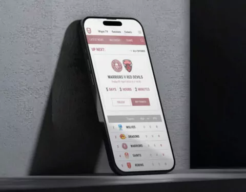 A smartphone displays a sports app; it shows upcoming game information, a countdown, and a league table. The phone rests on a ledge against a wall.